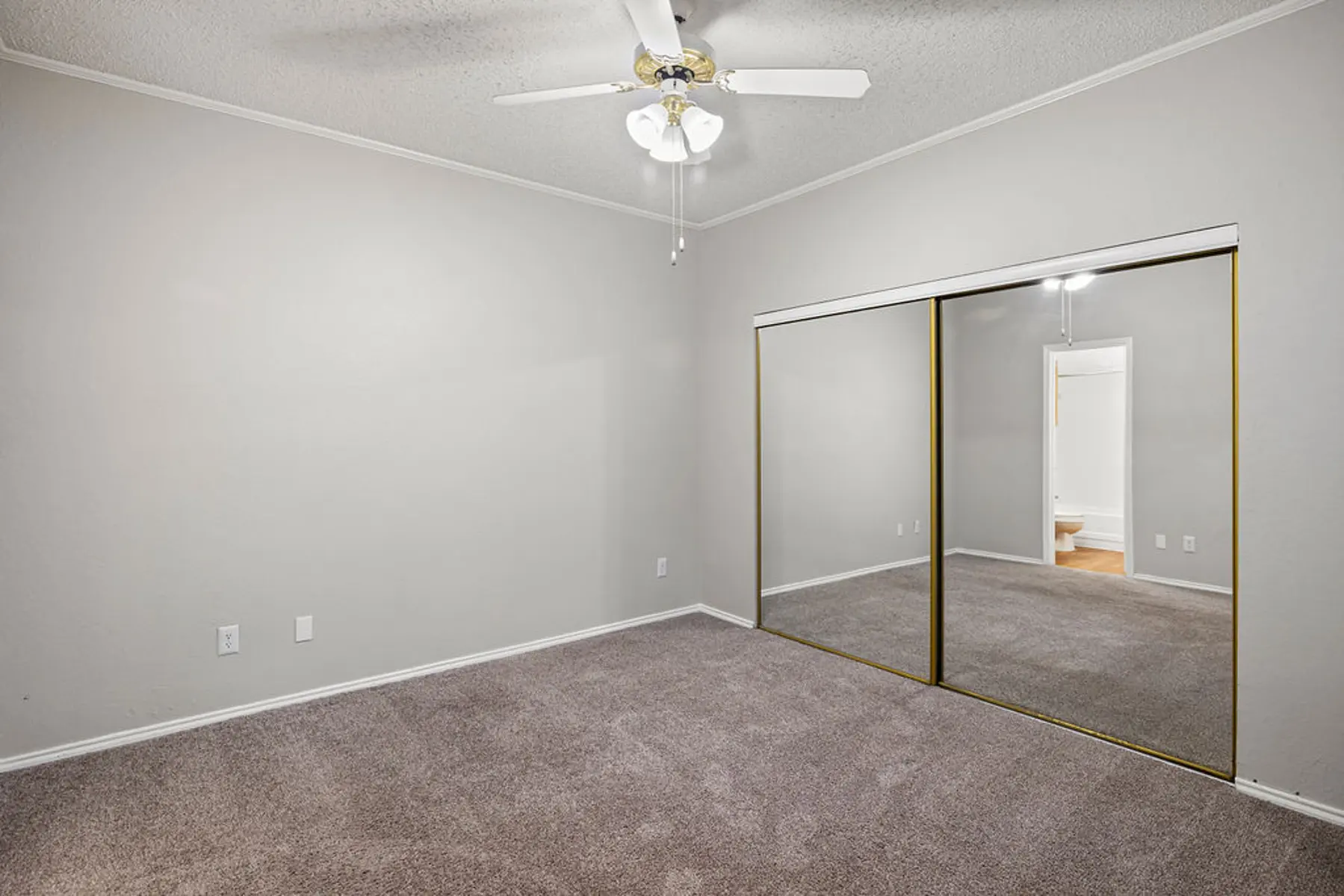 Vacant Bedroom with Ceiling Fan and Mirrored Closet A vacant bedroom featuring beige walls and carpet, equipped with a ceiling fan. A mirrored closet door is prominently displayed, reflecting light from the nearby room. The space is brightly lit and organized.
