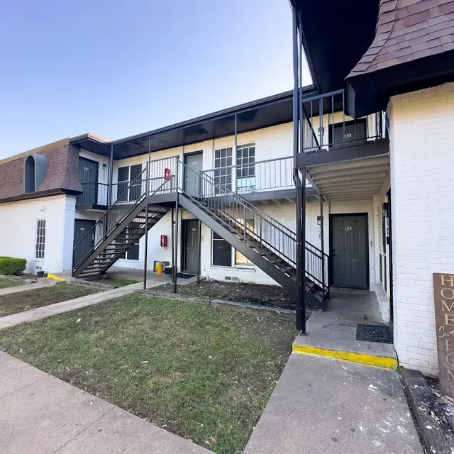 Front view of a two-story apartment complex with a staircase leading to upper units, grassy area in front, and a sign that reads 'HOME is where your story begins.'