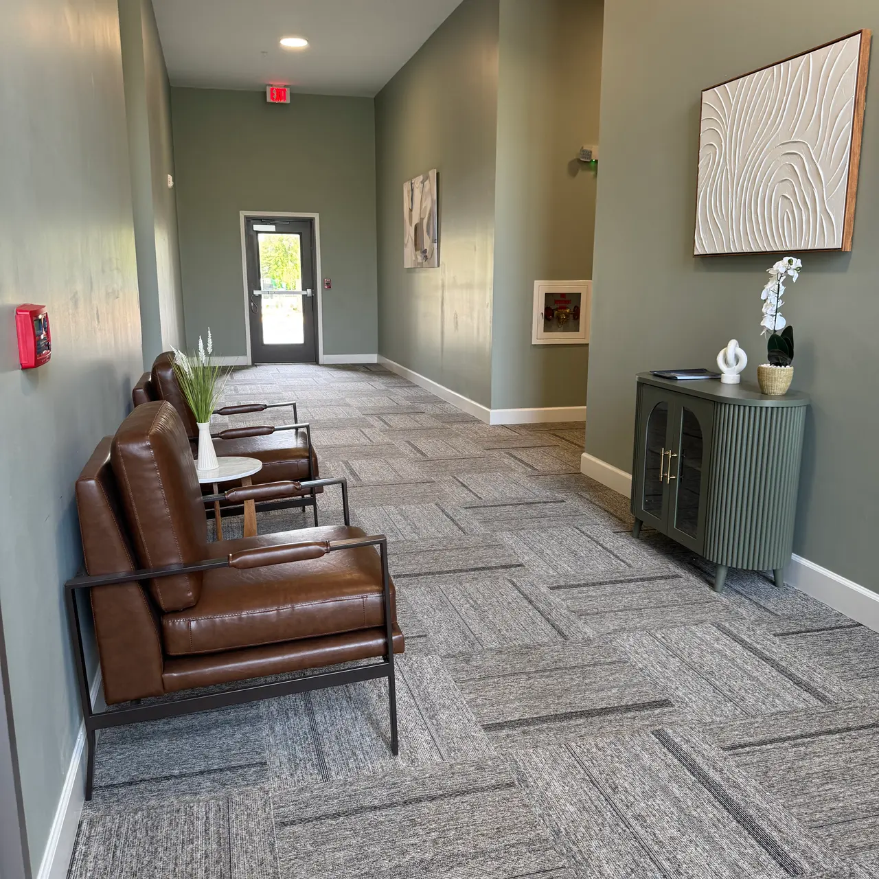 A modern hallway featuring two brown leather chairs, a small table, green walls, and a door at the end leading outside.