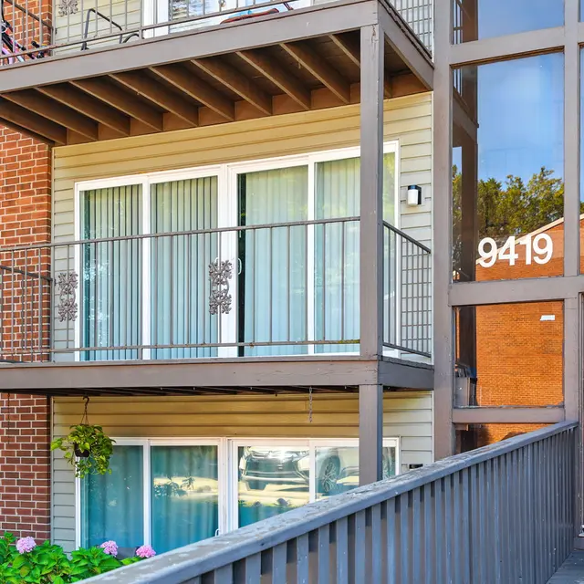 A modern three-story apartment building with a wooden balcony on the second level, featuring large windows with white curtains and flower pots. The building has a brick facade and a contemporary entrance with a glass door displaying the number 9419.
