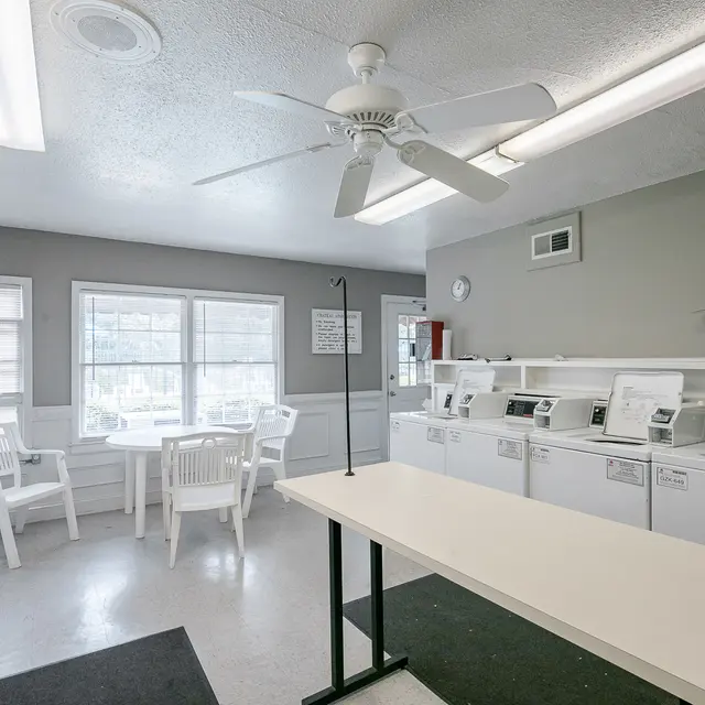 A clean, well-lit laundry room featuring multiple washing machines and dryers, a folding table, and seating area beside windows.