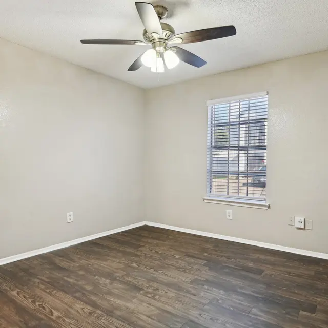 A vacant room with light brown walls and dark wooden flooring. There is a ceiling fan with light fixtures and a window with blinds, allowing natural light to enter the space.