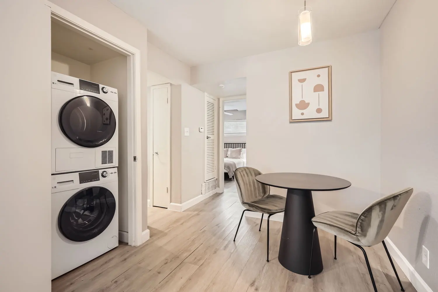 A modern laundry area featuring a stacked washer and dryer next to a small dining space with two chairs and a round table, leading into a hallway with a bedroom visible.