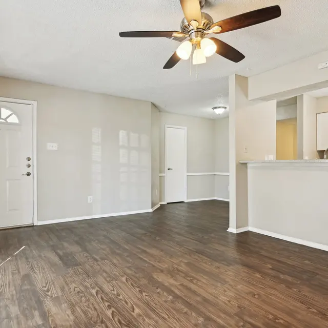 Interior of a living room featuring a ceiling fan, hardwood floors, and an entrance door. The space is well-lit with natural light and has a modern aesthetic.