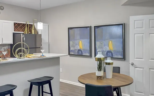 A modern kitchen and dining area featuring a sleek countertop, black bar stools, and two framed abstract paintings on the wall. There's a round wooden dining table with decorative vases and glasses placed atop it.