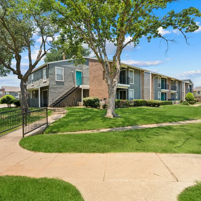 A well-maintained apartment complex surrounded by green grass and trees, featuring a concrete pathway, and a set of stairs leading to the second floor of one of the buildings.