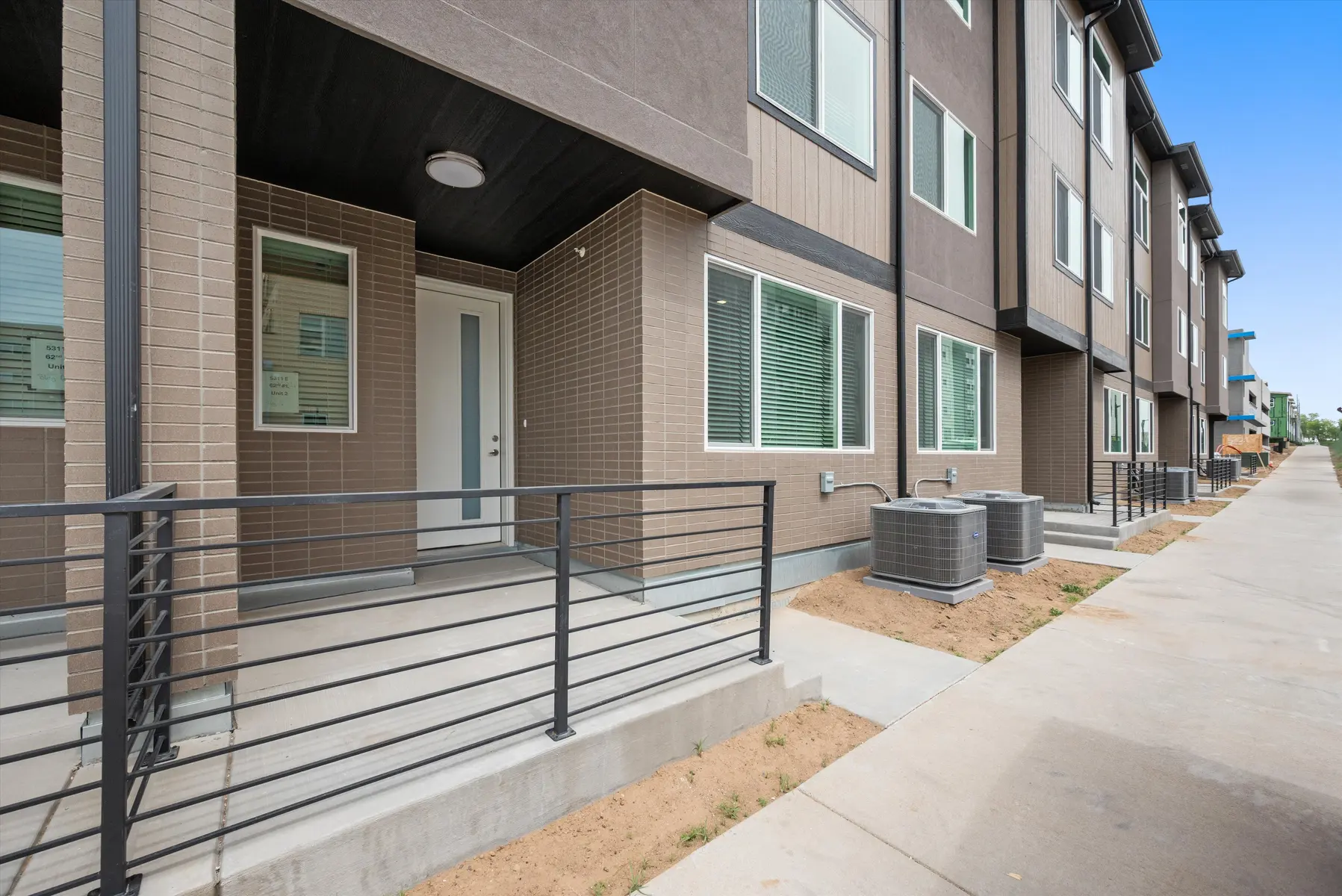 Modern Apartment Exterior View Exterior view of a modern apartment complex featuring beige and brown siding with a porch area and railings. The scene shows several air conditioning units and a walkway leading down the complex.