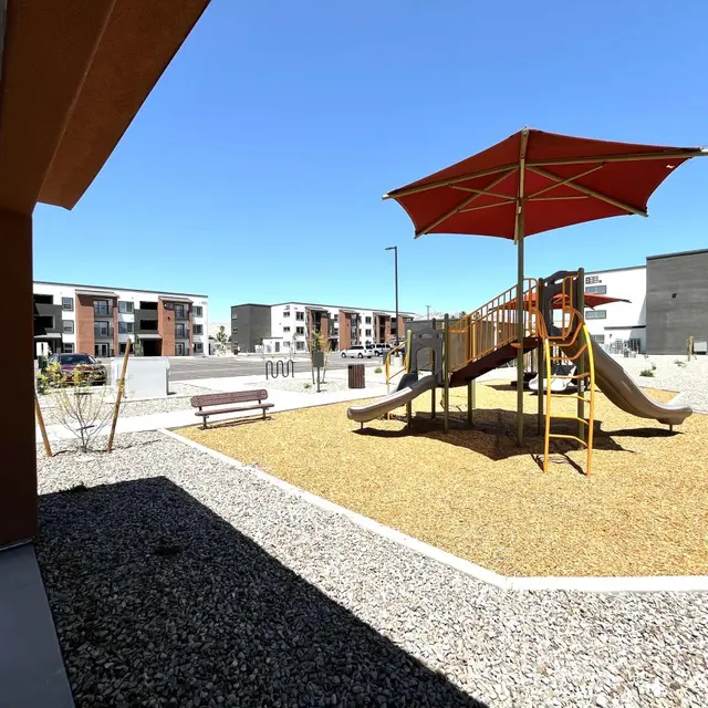 Children's Playground in Residential Area A playground featuring a wooden slide structure with a red canopy, surrounded by gravel and benches, and modern apartment buildings in the background under a clear blue sky.