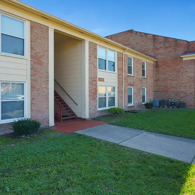 Exterior view of a two-story apartment building with a staircase, surrounded by grass and bushes under a clear blue sky.