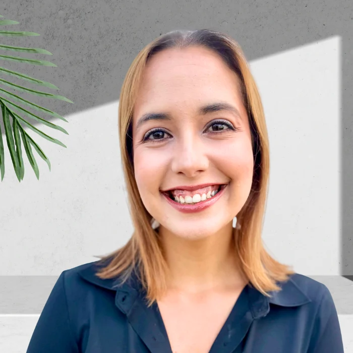 A smiling young woman stands against a minimalist background featuring a green plant and a gray wall. She has shoulder-length hair and is wearing a black top.