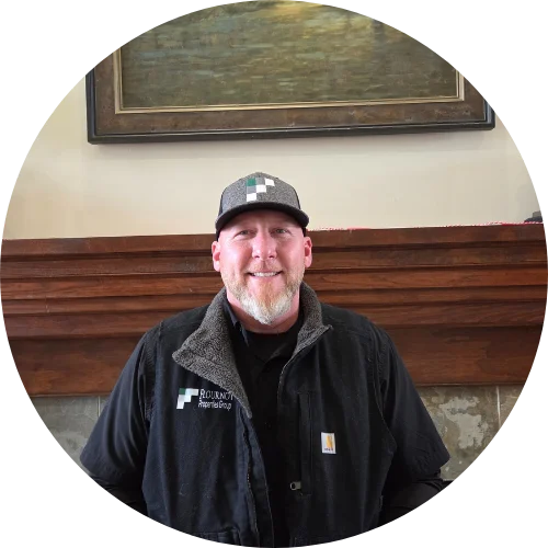 A smiling man in a black jacket and cap, standing in front of a wooden mantel.