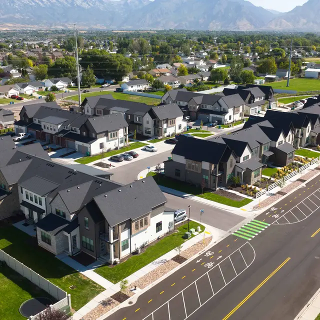 Aerial view of a new residential neighborhood with multiple houses. The houses feature modern architecture with dark roofs and landscaped yards. In the background, there are mountains and open green spaces.