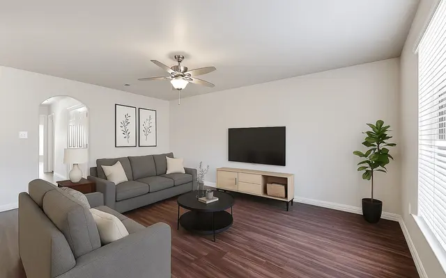 A modern living room featuring a gray sofa, a round black coffee table, and a wall-mounted TV. The walls are light-colored, and there are framed art pieces and a decorative plant in the corner.