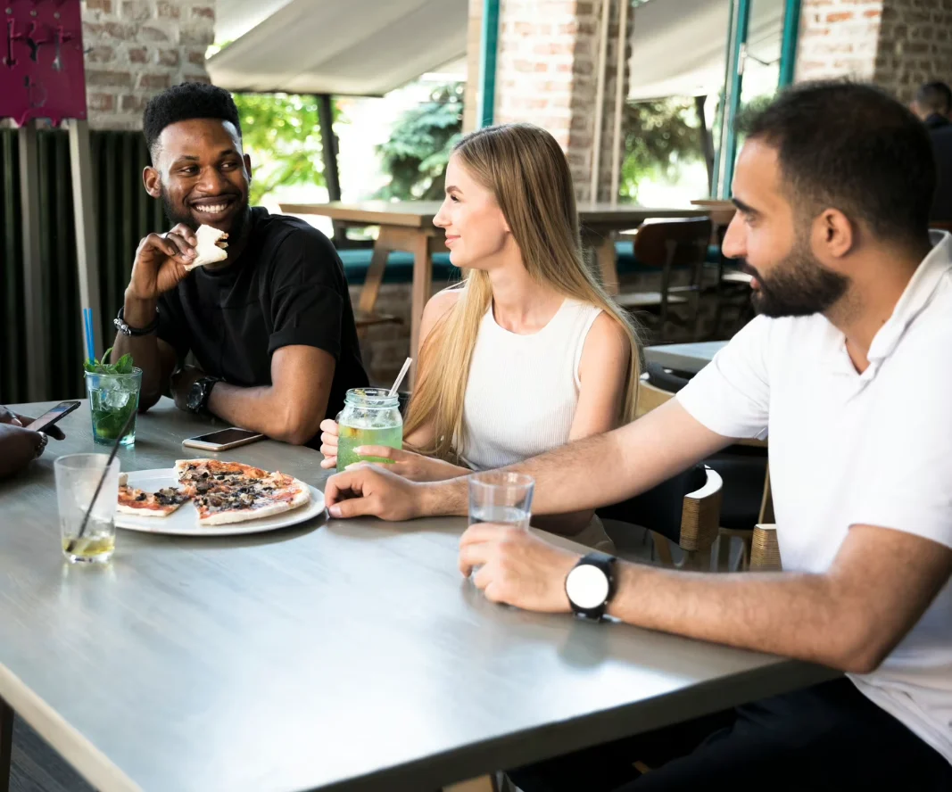 Casual Dining with Friends A group of three friends enjoying a meal together at a restaurant. They are seated at a table with drinks and food in front of them, smiling and engaged in conversation.