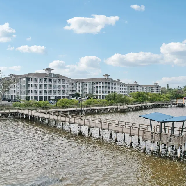 Scenic Pier View with Residential Building A wooden pier extending into a calm body of water, with a large multi-story residential building in the background under a blue sky with fluffy clouds.