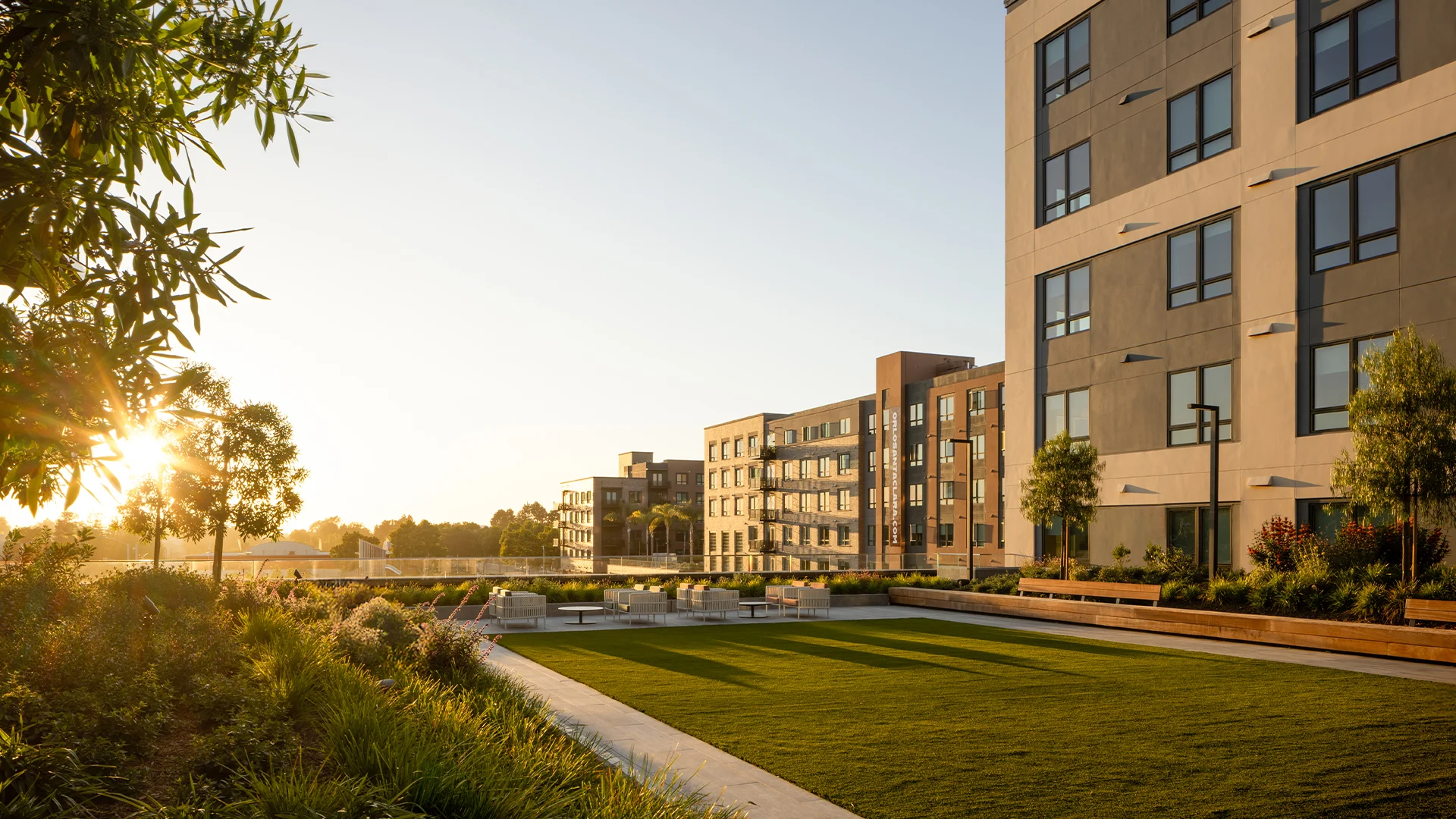 Modern Apartment Complex at Sunset A modern apartment complex with multiple buildings set against a sunset. The foreground features a neatly manicured lawn with lounge chairs, surrounded by greenery. The buildings display a contemporary architectural style.