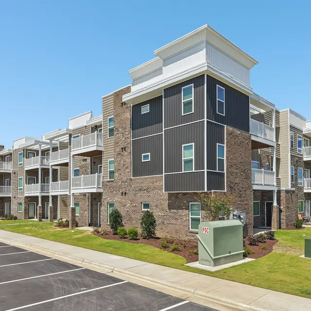A modern apartment building with a mix of brick and dark siding, featuring multiple balconies and a well-maintained front lawn. The parking area in front is paved with several spaces available.