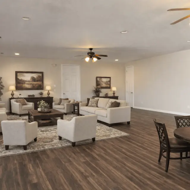 A spacious living room featuring comfortable seating arrangements with light-colored sofas and chairs, a center coffee table, end tables with decorative items, and a dining area with a round table and chairs. The room has wood flooring and large windows allowing natural light to brighten the space.