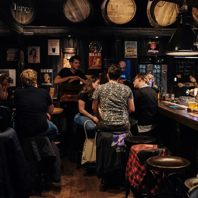 Hearsay Market Square A dimly lit pub interior with patrons seated at a wooden bar and tables. There are decorative beer barrels on the walls and a bartender serving drinks. Several people are engaged in conversation and enjoying beverages.
