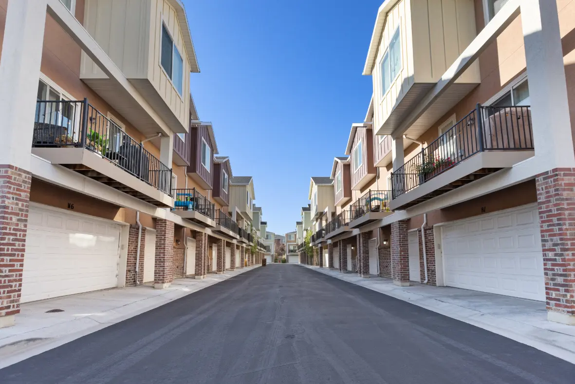 A street view of modern townhouses lined on either side, featuring garages and balconies, with a clear blue sky above.