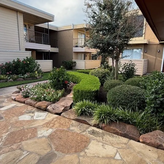 A landscaped courtyard with a stone path, surrounded by greenery and flowers, leading up to two-story buildings.