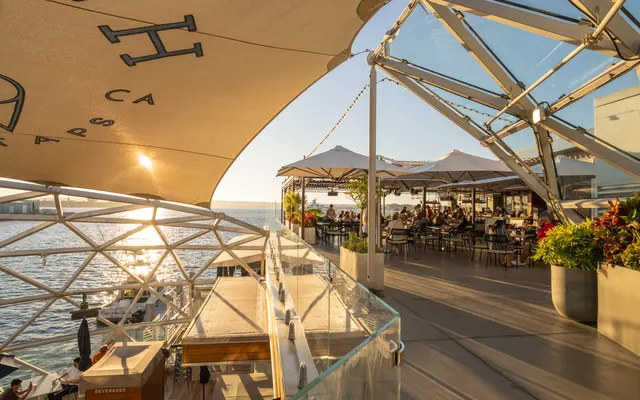 A waterfront dining area with a modern geodesic design. The sun is setting over the water, casting a warm glow on the scene. Outdoor seating is visible, with patrons enjoying their meals under large umbrellas.