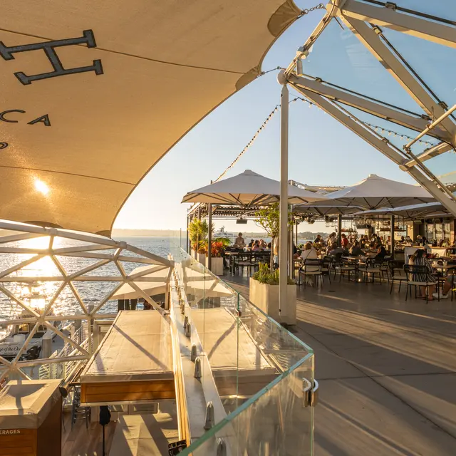 A waterfront dining area with a modern geodesic design. The sun is setting over the water, casting a warm glow on the scene. Outdoor seating is visible, with patrons enjoying their meals under large umbrellas.