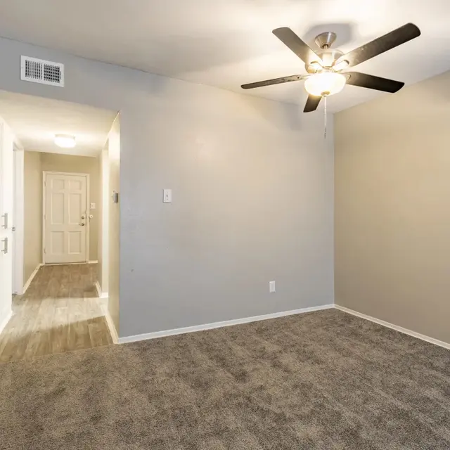 A spacious empty room featuring a ceiling fan and light, with beige walls and beige carpeting.