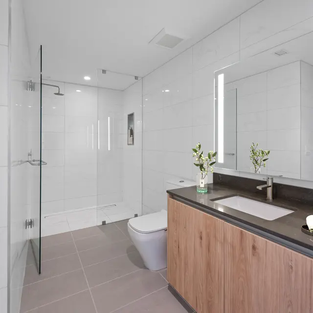 A modern bathroom featuring large white tiles, a walk-in shower with glass doors, a wooden vanity with a dark countertop, and elegant lighting fixtures.