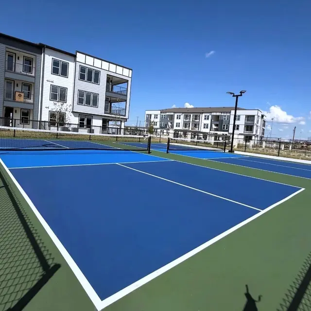 A modern residential complex with two blue tennis courts surrounded by green surfaces and appartment buildings in the background under a clear blue sky.