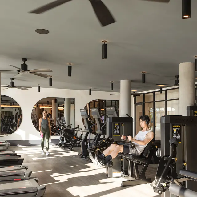 A modern fitness gym featuring various exercise equipment and several people engaged in workouts. The space has large windows allowing natural light, and the decor includes sleek lines and contemporary design elements.