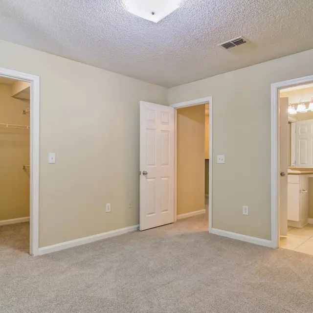 A spacious bedroom featuring a light brown carpet, two doors leading to separate rooms, and a doorway with a mirror that leads to a bathroom. The walls are painted a soft beige color. A closet is visible on the left side with a wire rack.