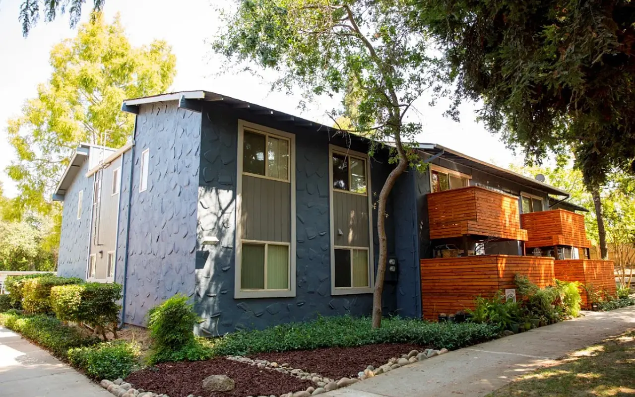 A modern apartment building featuring a blue exterior, large windows, and wooden balconies, surrounded by landscaped greenery.