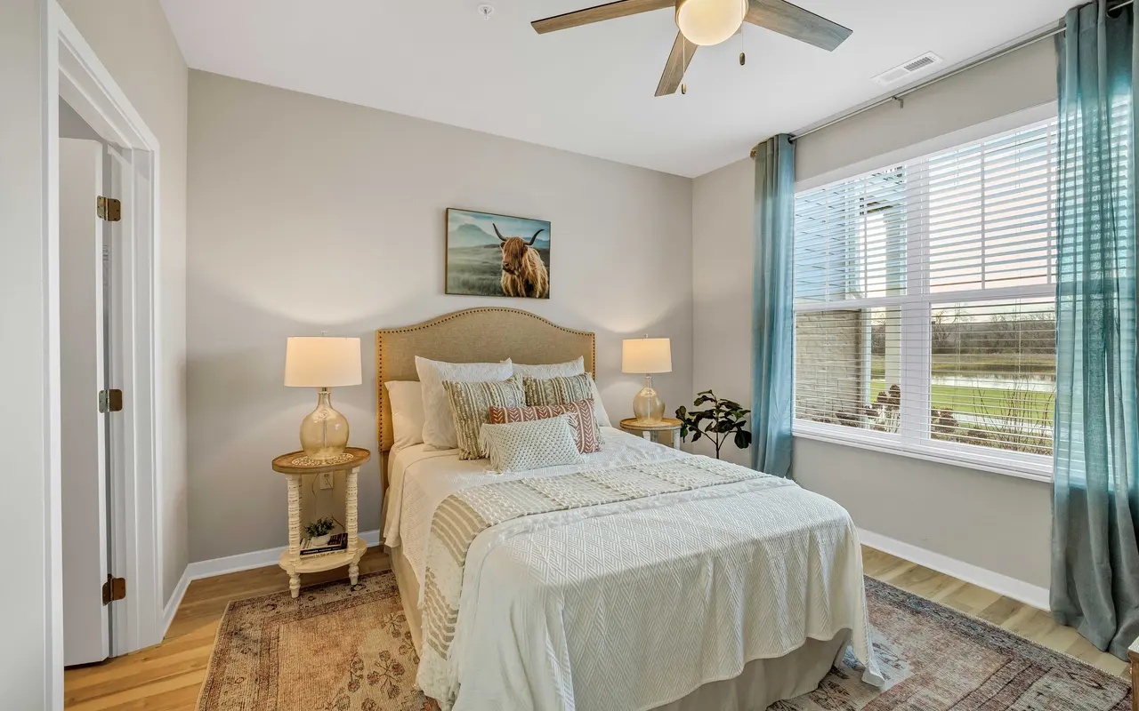 A cozy bedroom featuring a light-colored bedspread, decorative pillows, and two bedside lamps. The room has a window with white blinds, curtains, and a painting of an animal on the wall. Wooden flooring and a decorative rug enhance the inviting atmosphere.