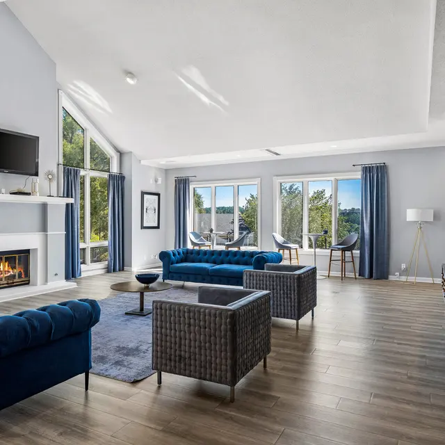 A spacious modern living room featuring blue furniture and a fireplace, with large windows letting in natural light and a view of greenery outside.