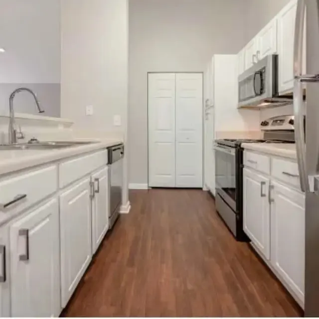 A modern kitchen featuring white cabinets, stainless steel appliances, and a wooden floor. The space is bright with natural light, showcasing sleek countertops and a sink.
