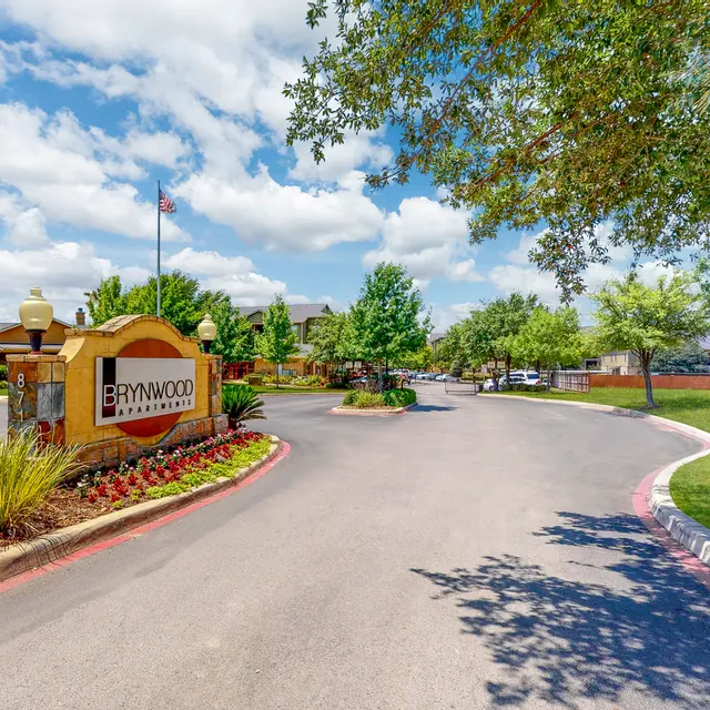 A welcoming entrance to Brynwood community featuring a sign, landscaped gardens, and a tree-lined pathway under a partly cloudy sky.