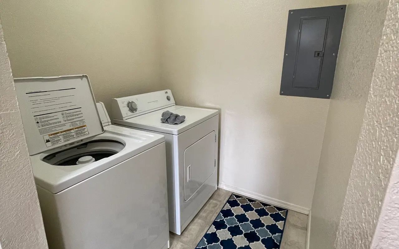A compact laundry room featuring a washing machine and dryer, with a patterned rug on the floor and a grey electrical panel on the wall.