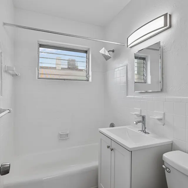 A bright and modern bathroom featuring a bathtub, sink with cabinet, and a small window for natural light. The walls are painted white, and there is a mirror above the sink.