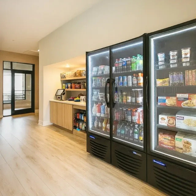 A modern kitchenette featuring a glass fridge filled with drinks and snacks, alongside a wooden shelf stocked with various food items. The floor has light wooden planks and the overall design is minimalistic and bright.