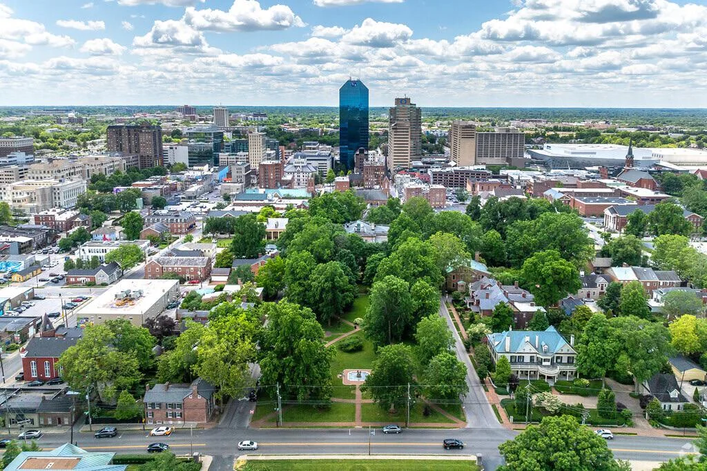 Urban Landscape with Greenery Aerial view of a city with a mix of modern skyscrapers and historic buildings, surrounded by lush greenery and open spaces under a partly cloudy sky.