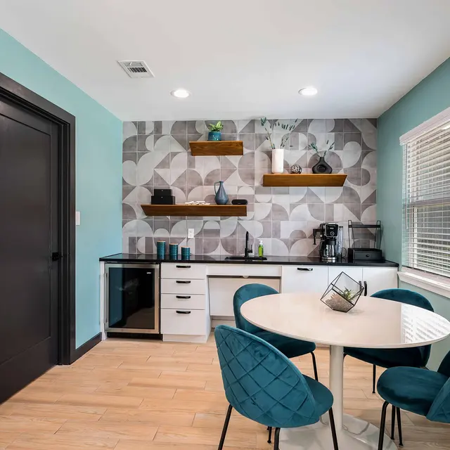 A modern kitchen and dining area featuring a round dining table with teal chairs, a black and white patterned wall, open shelving with decor items, and a black kitchen counter with appliances.