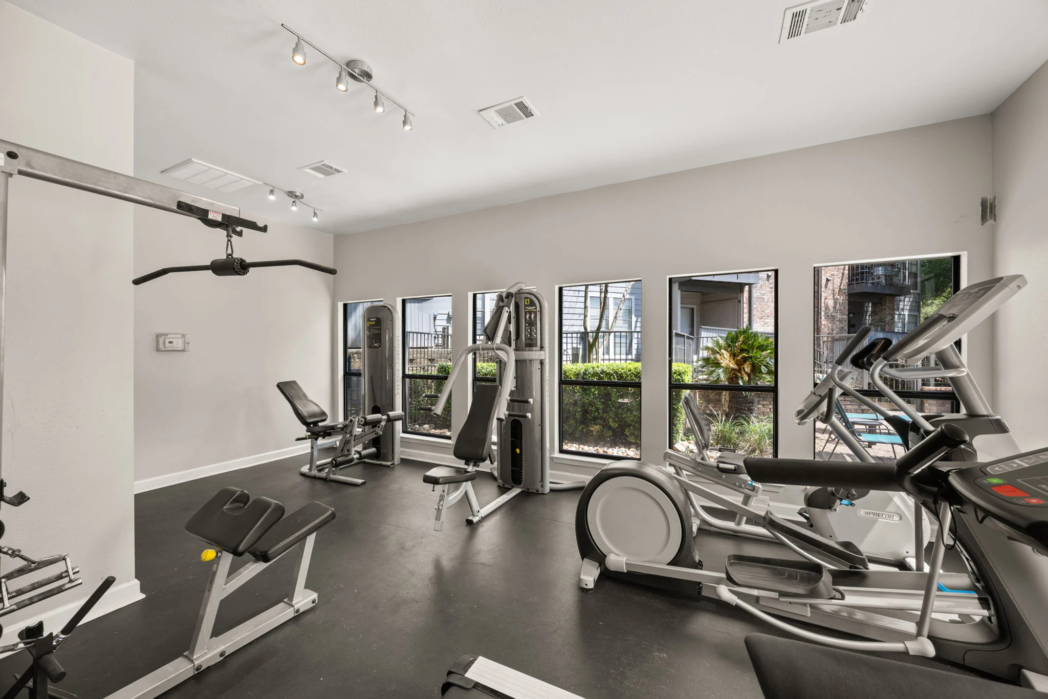 A modern fitness room featuring various exercise equipment including a treadmill, elliptical, stationary bike, and multi-gym weights. Large windows allow natural light and a view of greenery outside.