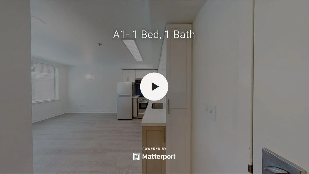 Interior view of a modern apartment with an open floor plan, featuring a kitchen area on the left and a spacious living area with large windows.