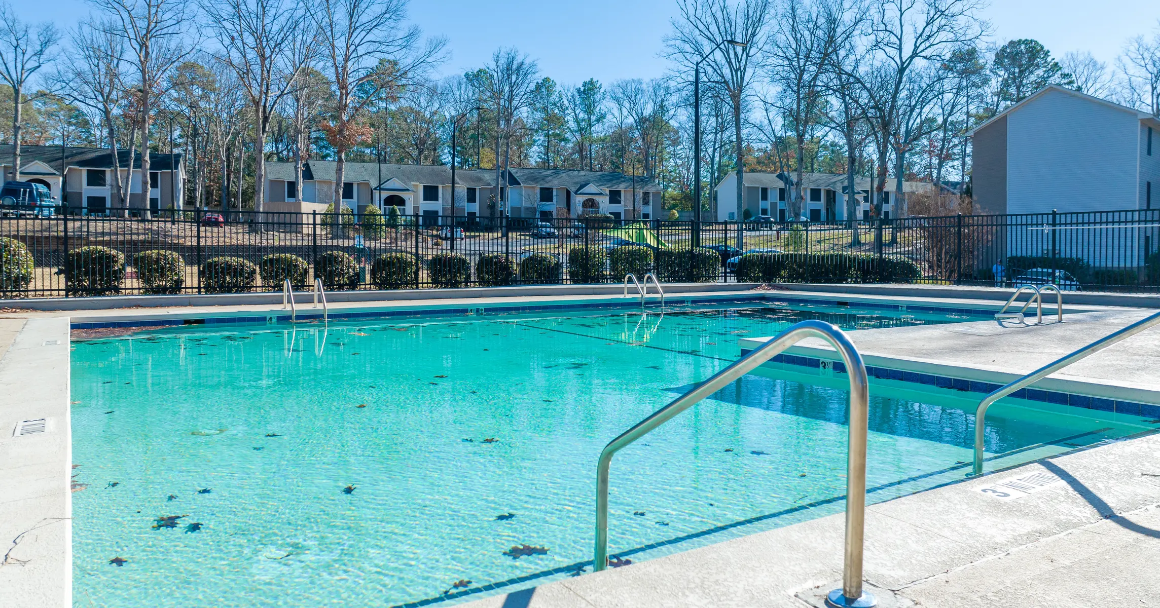 A clear swimming pool surrounded by a fenced area, with a view of apartment buildings in the background and bare trees nearby.