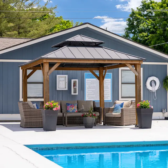 A poolside area featuring a wooden gazebo with comfortable seating and colorful flower pots, beside a clear blue swimming pool.