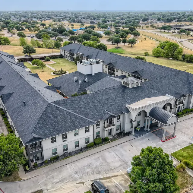 Aerial view of a two-story apartment complex with a flat roof, surrounded by green trees and open land. The complex features a central entrance with an awning.