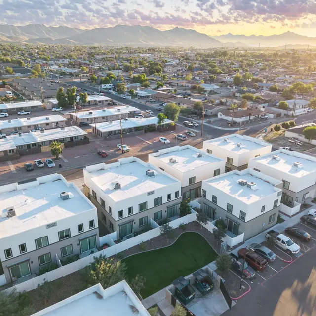 Aerial View of Suburban Residential Area Aerial view of a suburban residential area featuring modern white-roofed homes and surrounding parking lots, with mountains in the background. The neighborhood shows a mix of single-family homes and multi-unit buildings, bathed in warm afternoon light.