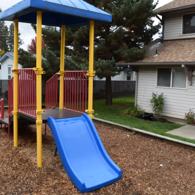 Playground Structure with Slide A small playground structure with a blue slide, red railings, and a blue roof, surrounded by a gravel area and a grassy lawn. In the background, there is a residential building and some trees.