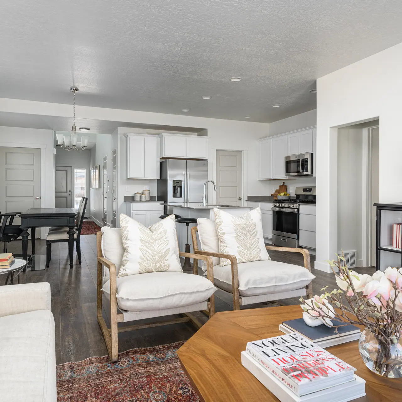 A modern living room featuring two armchairs and a sofa with decorative pillows, a wooden coffee table, and a vase with flowers. The space includes a dining area in the background, modern kitchen appliances, and light-colored walls.